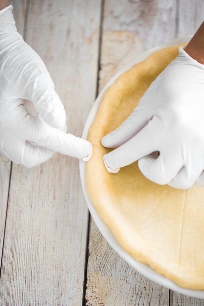 Hands in gloves crimping the edges of an unbaked meat pie to seal the crust