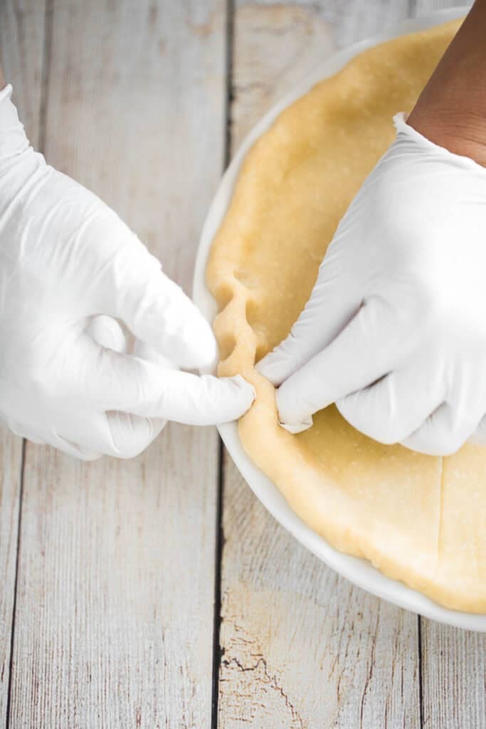 Hands in gloves pinching and crimping the edge of an unbaked pie crust