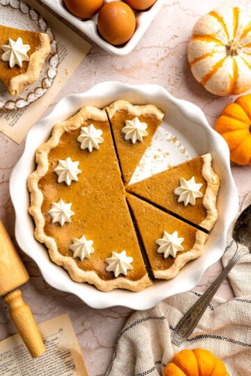 Homemade pumpkin pie in a white fluted pie dish, topped with whipped cream rosettes, with slices cut and one slice removed.