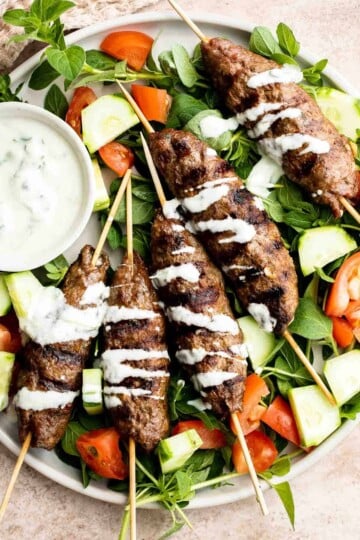 Beef Kofta Kebabs served over a fresh cucumber and tomato salad with creamy yogurt sauce drizzled on top.