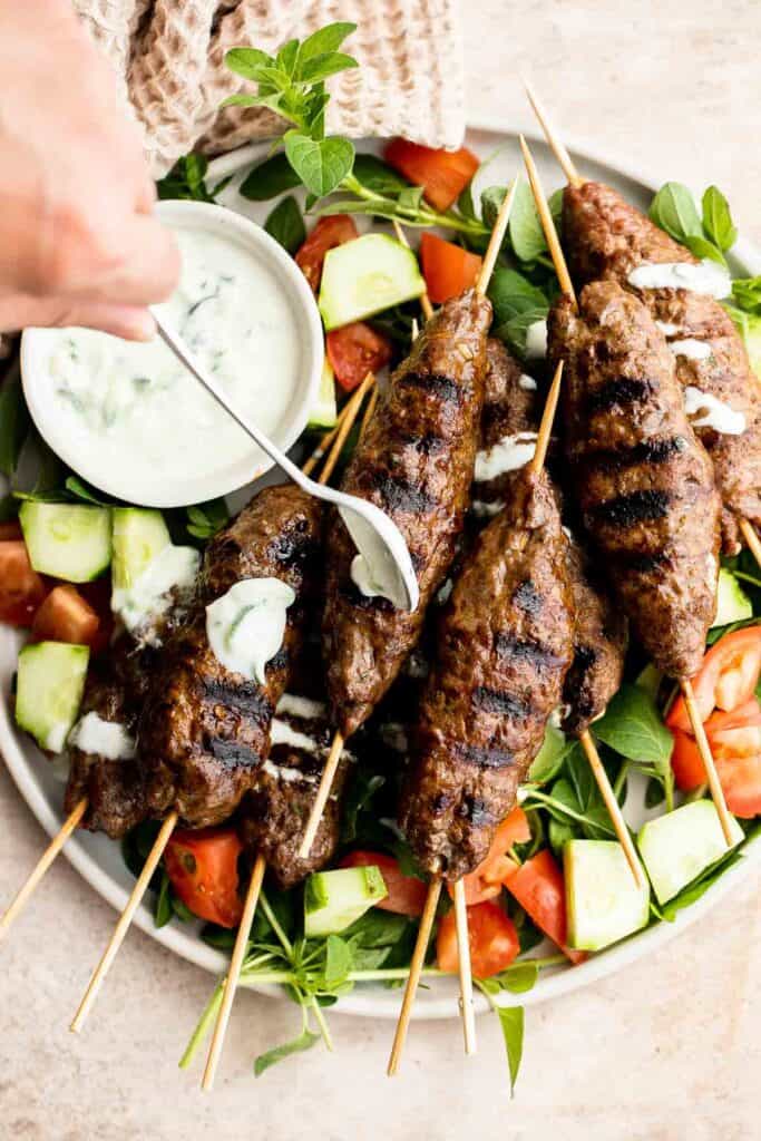 Hand drizzling creamy yogurt sauce over Beef Kofta Kebabs served with cucumber and tomato salad.