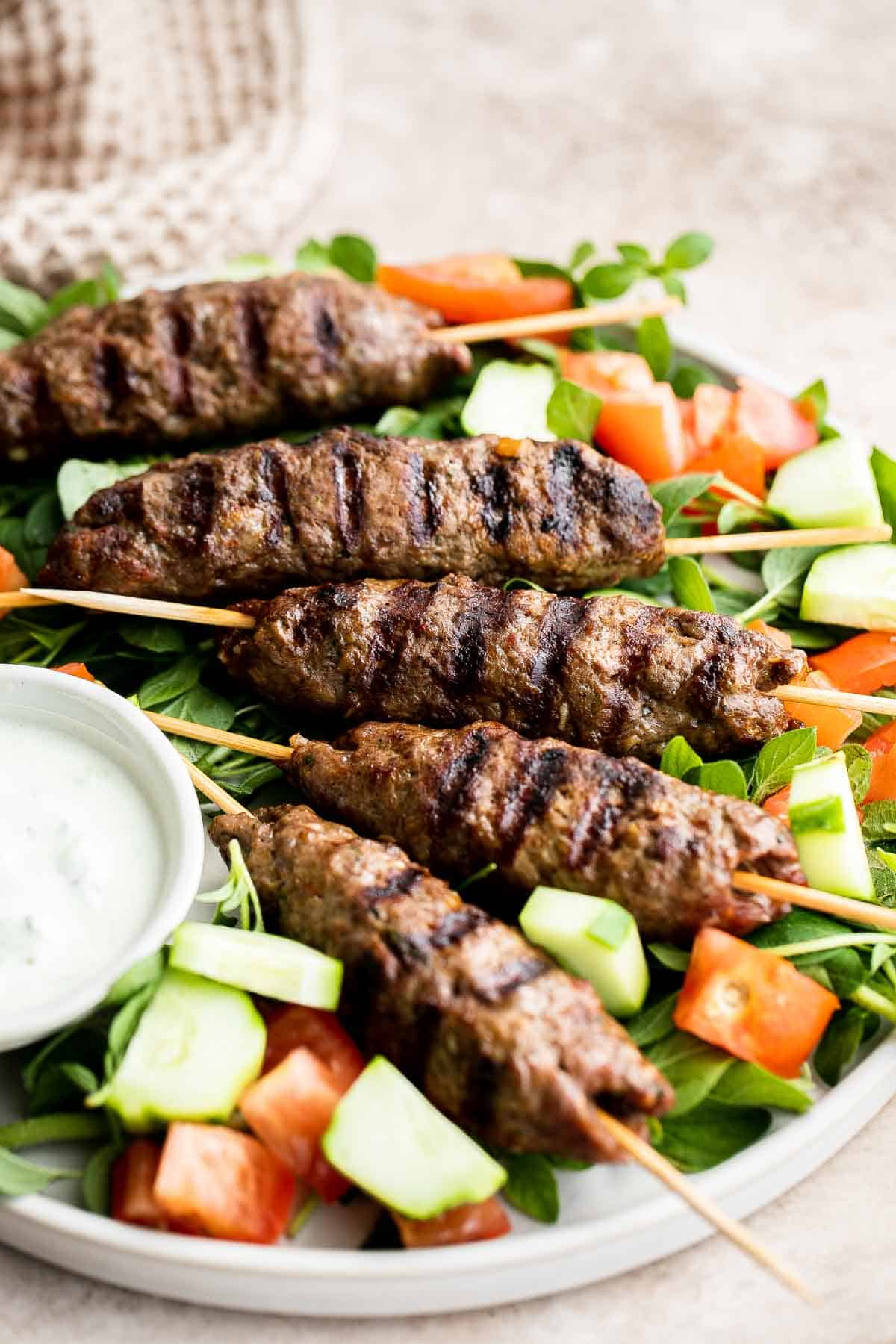 Close up of grilled Beef Kofta Kebabs served with cucumber, tomato, and fresh greens.