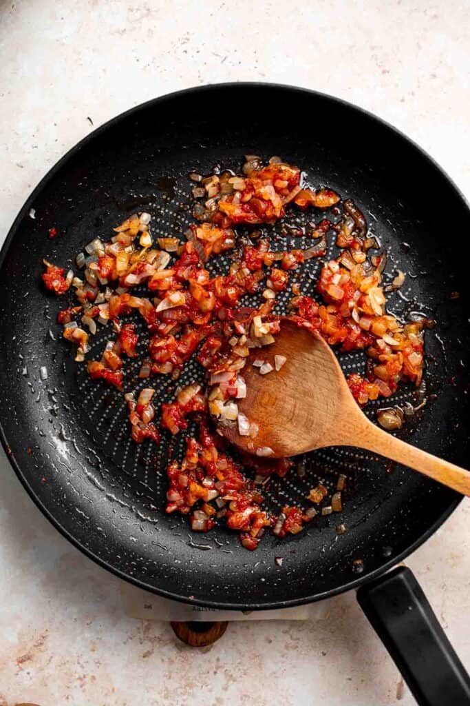 Tomato paste and spices being stirred into sautéed onion and garlic in a skillet