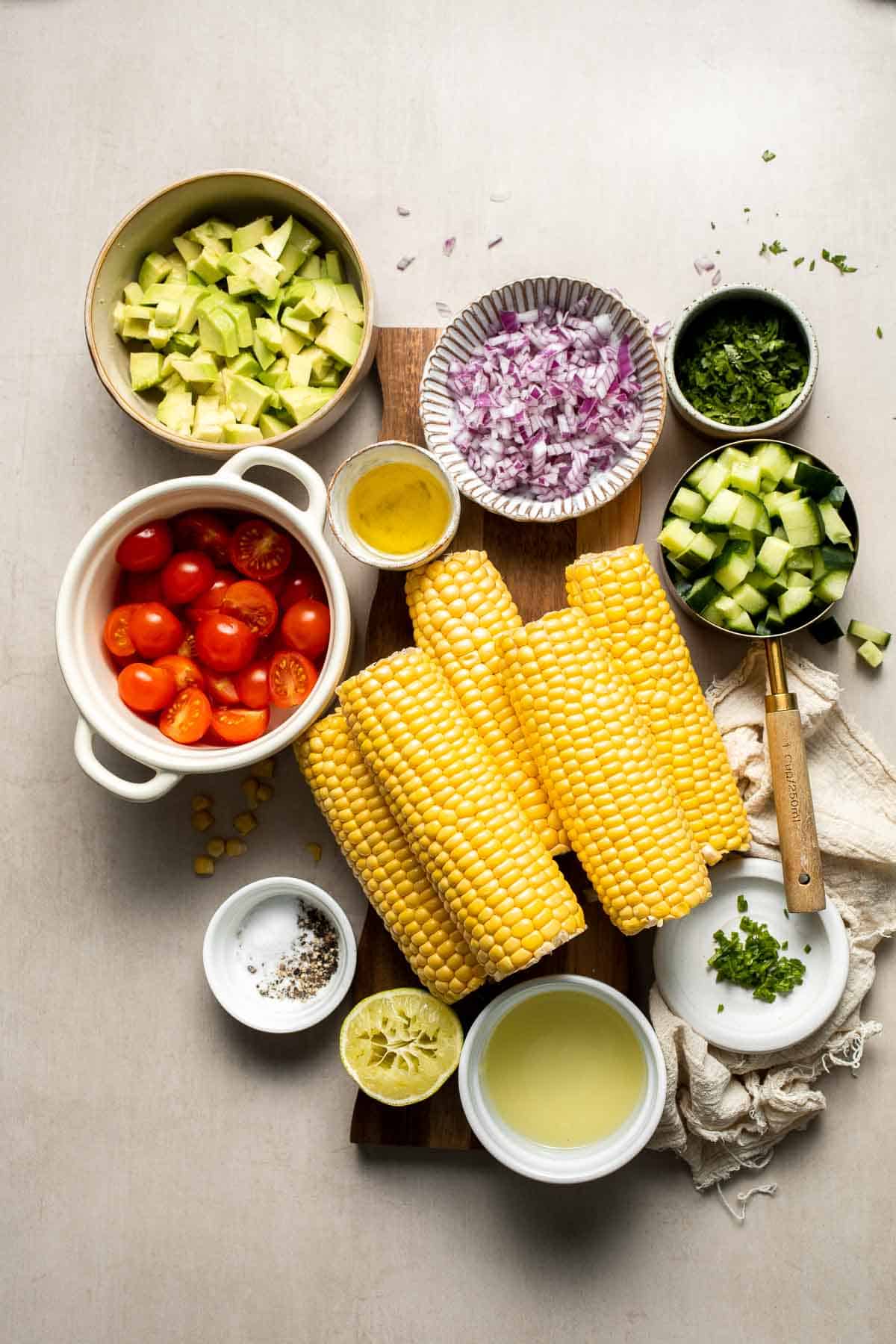 Ingredients for avocado corn salad including fresh corn, avocado, cherry tomatoes, cucumber, red onion, cilantro, lime, and olive oil.