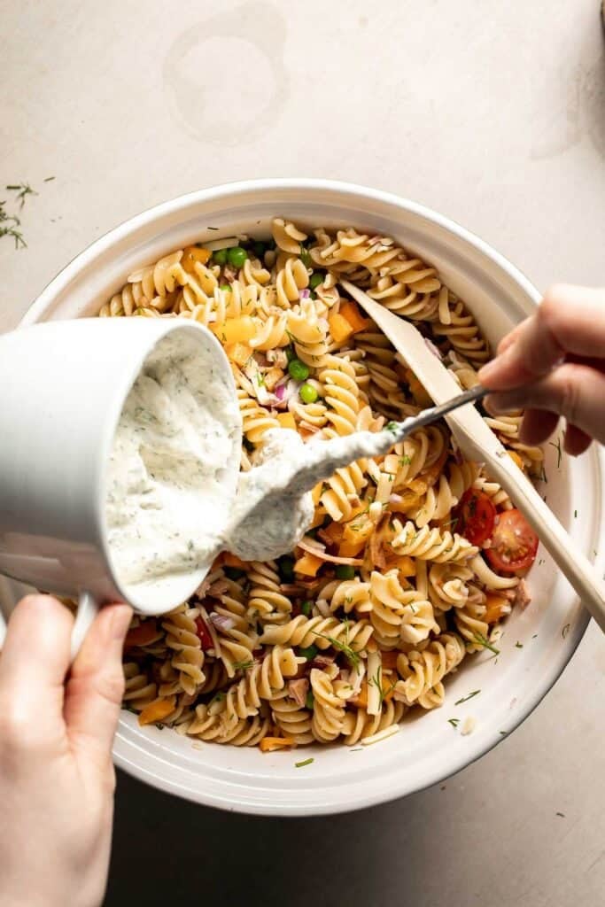 Creamy ranch dressing being poured over pasta salad ingredients.
