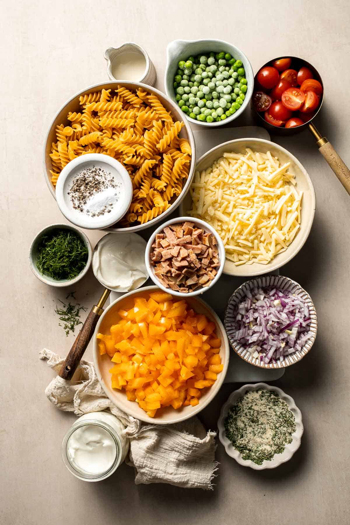 Ingredients for bacon ranch pasta salad including rotini pasta, bacon, cheddar cheese, peas, tomatoes, bell pepper, red onion, ranch dressing ingredients, and fresh herbs.