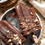 Homemade fudgesicles drizzled with melted chocolate and topped with chopped nuts on a serving platter.