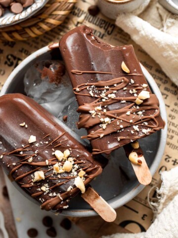 Homemade fudgesicles drizzled with melted chocolate and topped with chopped nuts on a serving platter.