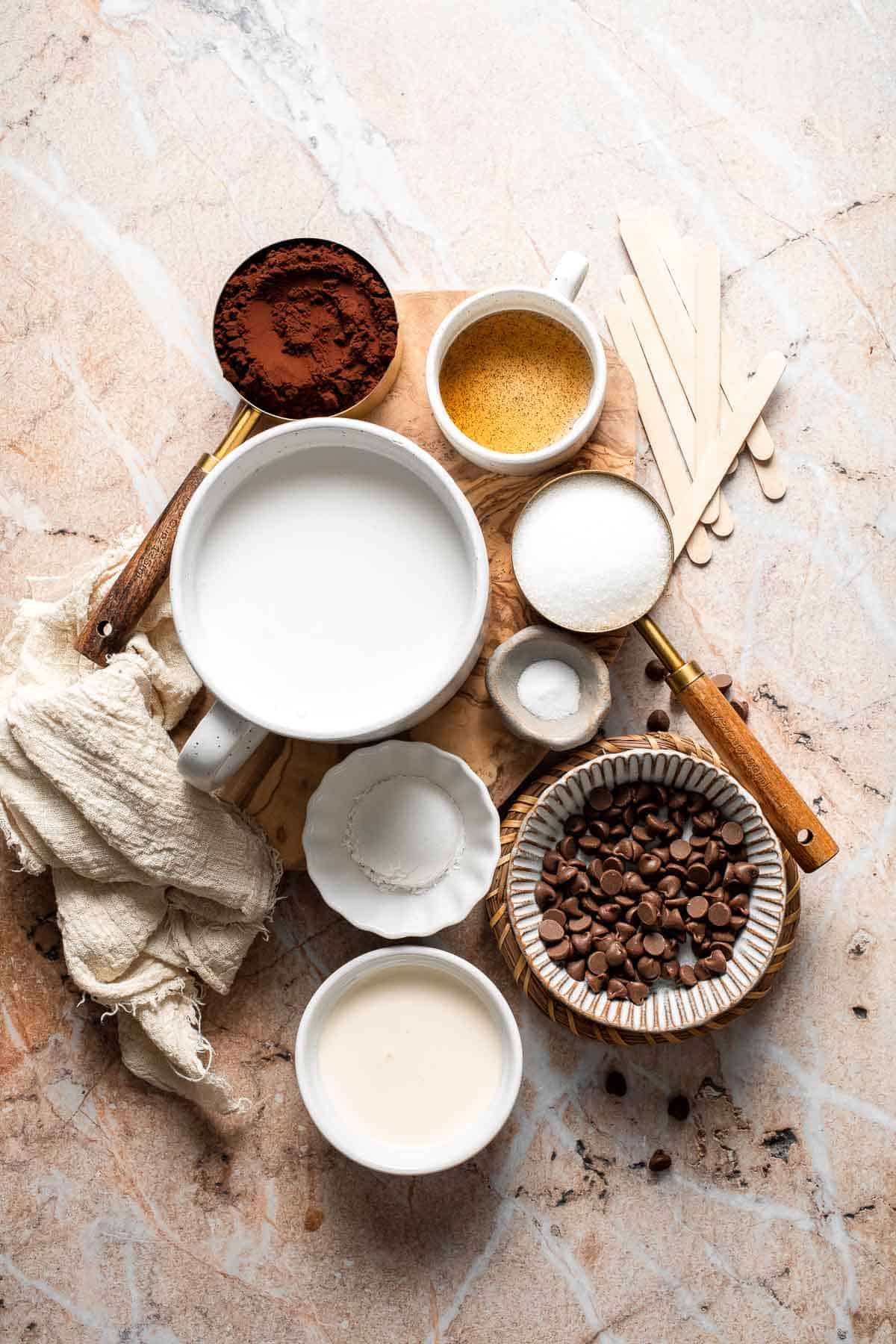 Ingredients for homemade fudgesicles including cocoa powder, milk, cream, sugar, chocolate chips, vanilla, and popsicle sticks.