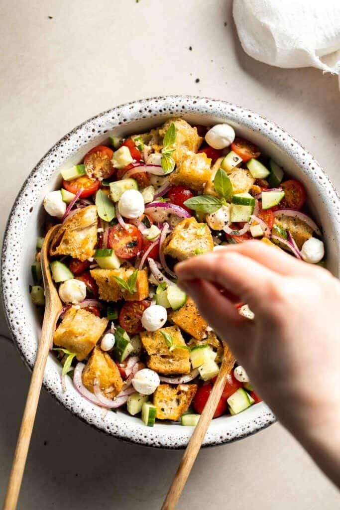 Hand sprinkling fresh basil over panzanella salad with toasted bread, tomatoes, cucumber, and mozzarella