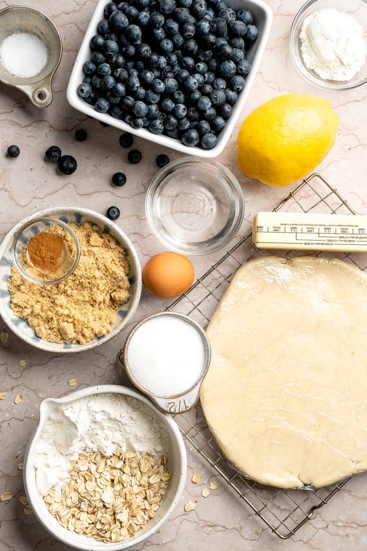 Ingredients for blueberry crumble pie including fresh blueberries, pie crust, oats, butter, lemon, sugar, and cinnamon.