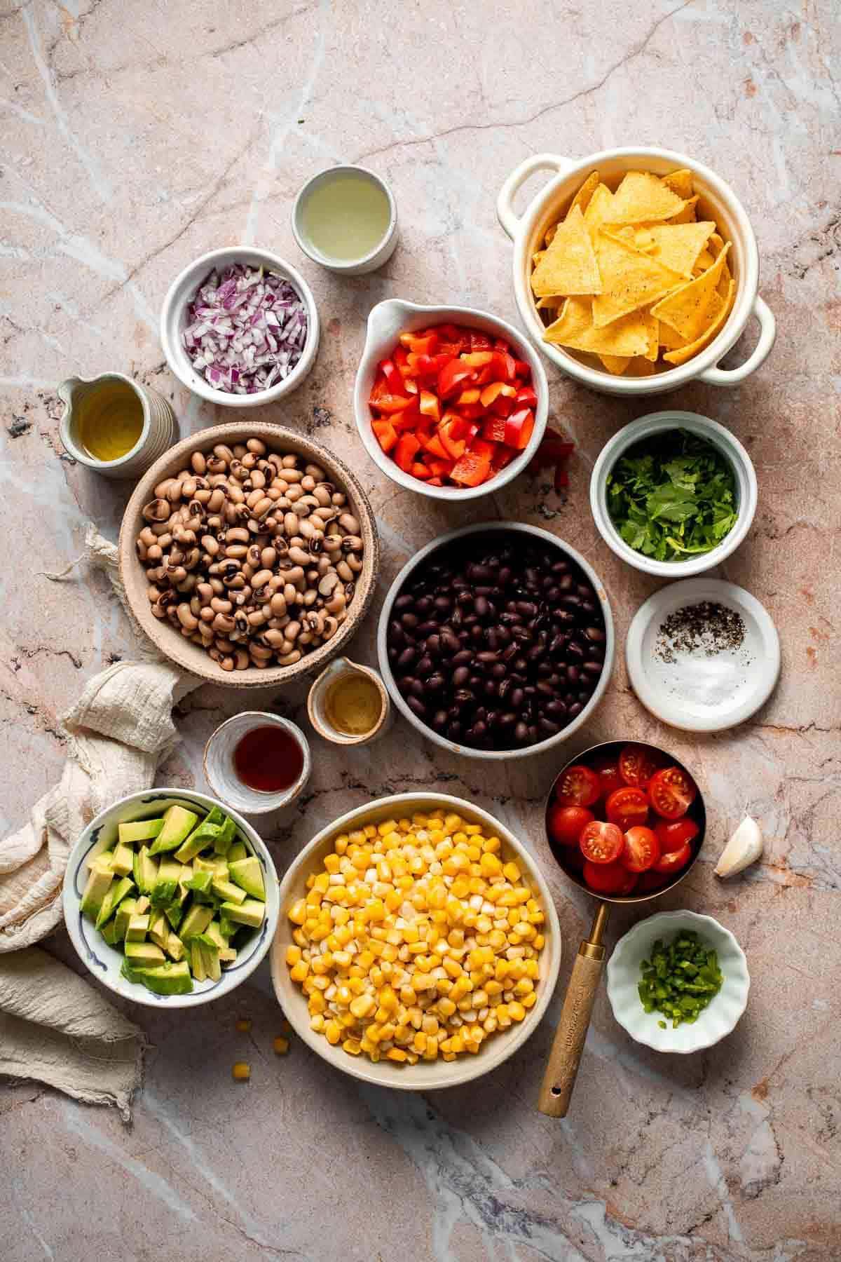 Ingredients for homemade Cowboy Caviar arranged in small bowls, including black beans, black-eyed peas, corn, avocado, tomatoes, peppers, onion, and cilantro.