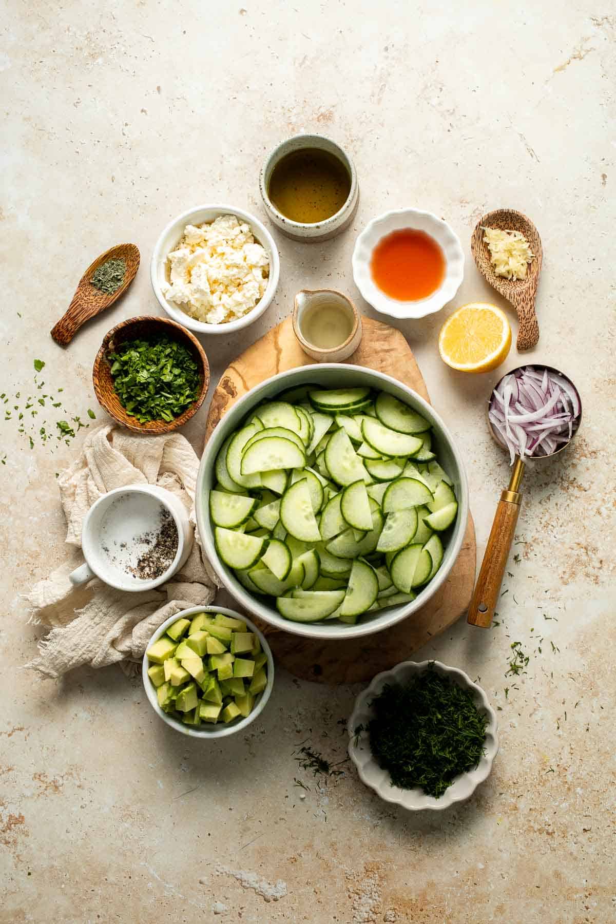 Ingredients for cucumber feta salad including cucumbers, avocado, feta cheese, red onion, fresh herbs, and dressing ingredients.