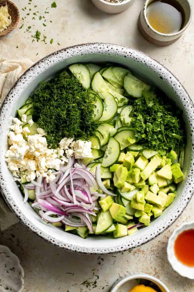 Sliced cucumbers, avocado, feta cheese, red onion, dill, and parsley in a large mixing bowl.