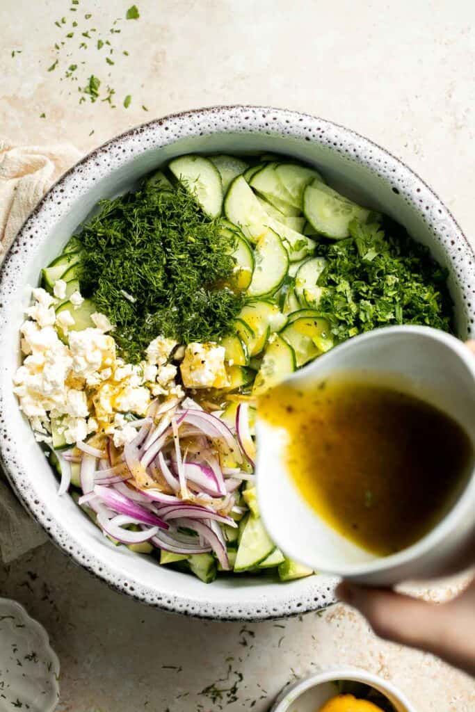 Pouring lemon vinaigrette over cucumber, avocado, feta, and fresh herbs.
