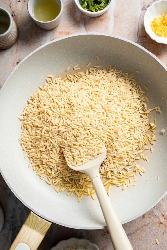 Dry orzo being stirred with garlic in a skillet before toasting.