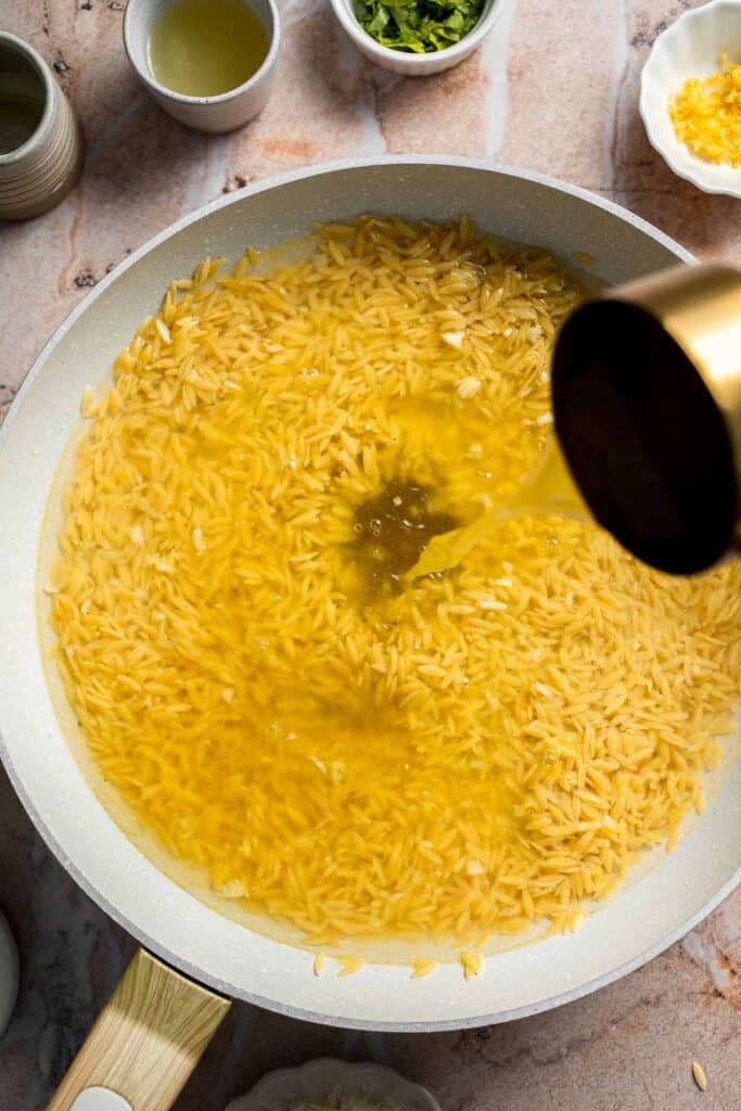 Pouring vegetable broth into toasted orzo in a skillet.