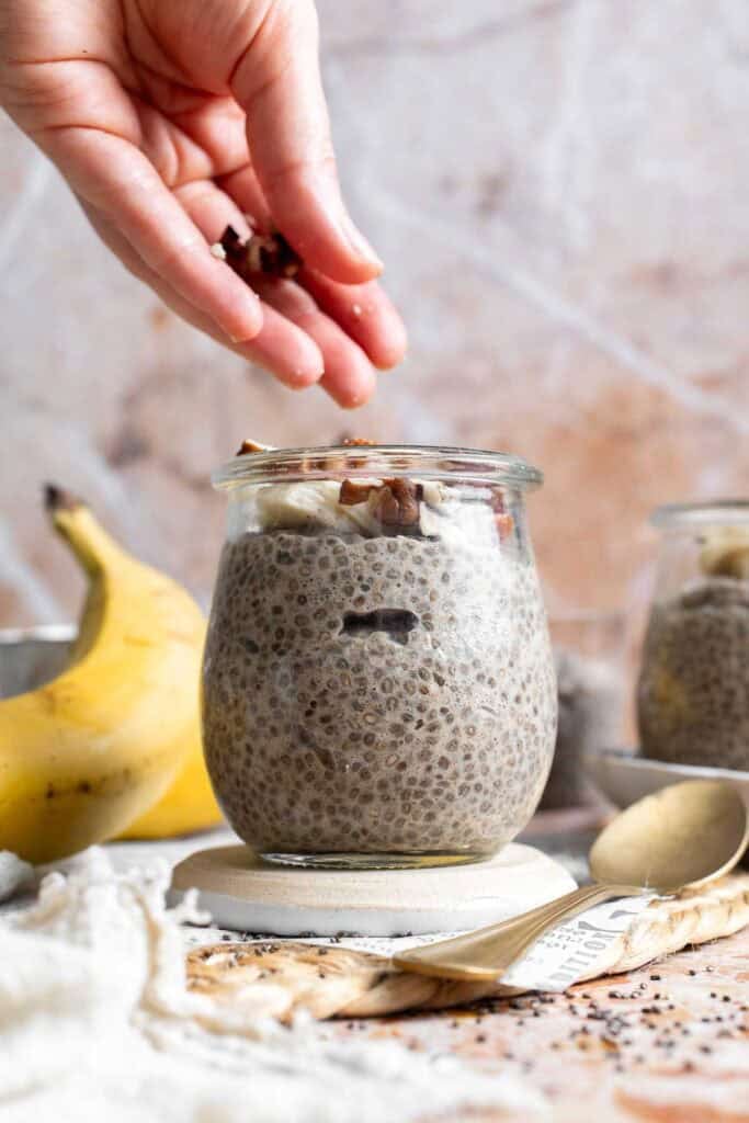 A hand adding chopped nuts to the top of banana chia pudding