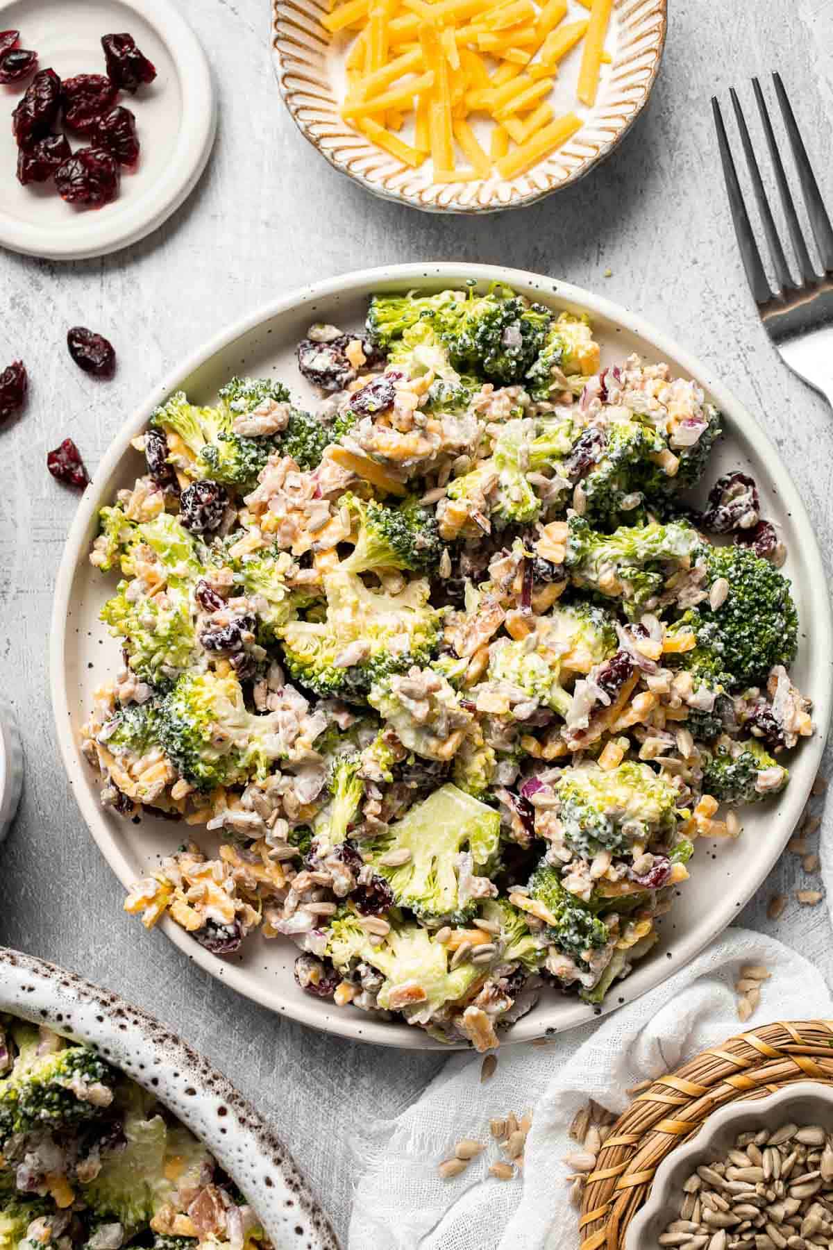 Broccoli Salad served in a plate with cheddar cheese and dried cranberries on the side.