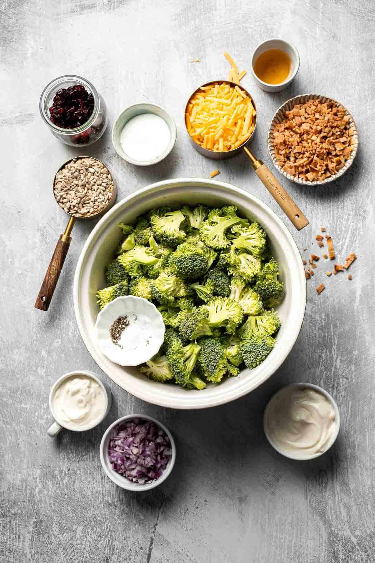 Ingredients for Broccoli Salad including broccoli, bacon, cheddar cheese, dried cranberries, sunflower seeds, red onion, and dressing ingredients.