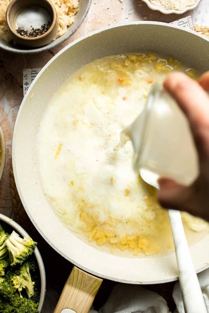 Milk being poured into a skillet with broth and roux, stirred with a spatula