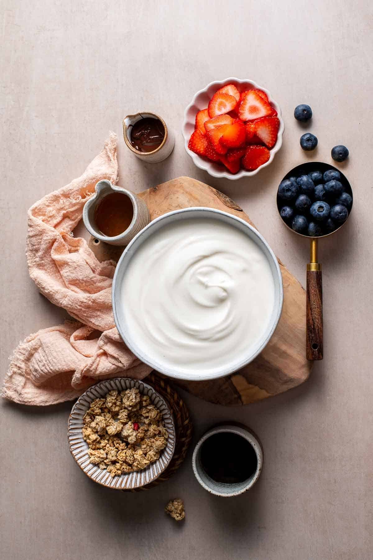 Ingredients for frozen yogurt bark including Greek yogurt, strawberries, blueberries, and granola arranged on a table