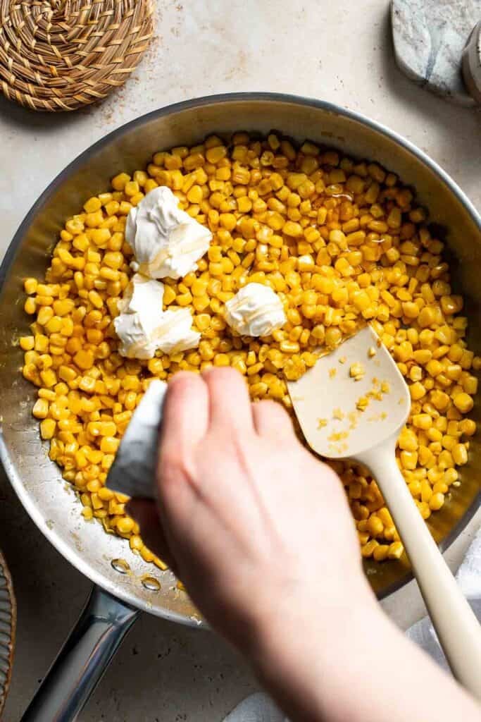 Cubed cream cheese being added to cooked corn in a skillet