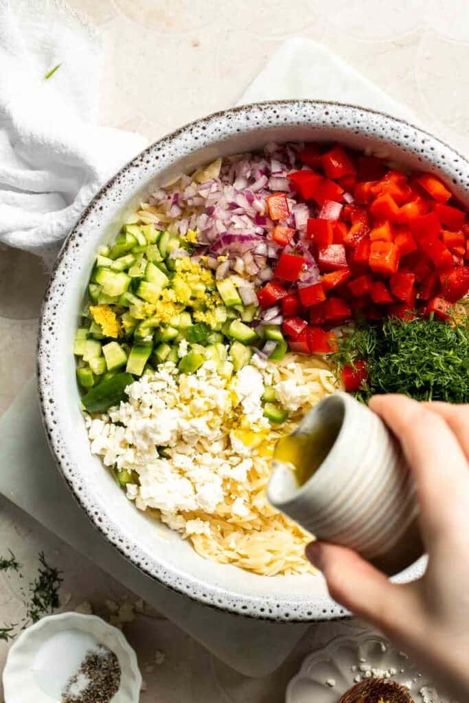 Adding olive oil to cooked orzo with cucumber, red pepper, red onion, feta, and dill for Lemon Orzo Salad.