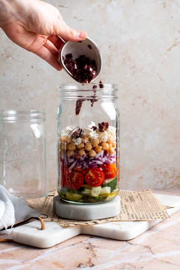 Black beans being poured into a mason jar layered with Mediterranean salad ingredients