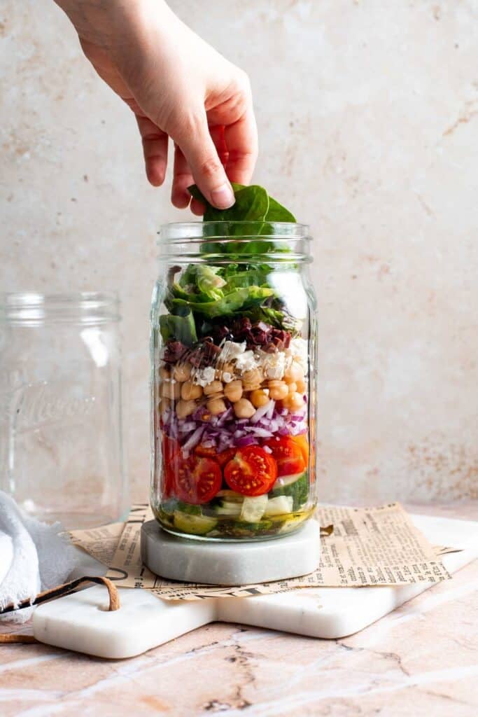 Mixed greens being added to the top of a layered Mediterranean mason jar salad