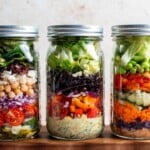 Three mason jar salads lined up showing colorful layered ingredients, including Mediterranean, Southwest, and Asian peanut variations