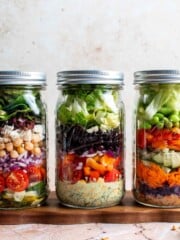 Three mason jar salads lined up showing colorful layered ingredients, including Mediterranean, Southwest, and Asian peanut variations