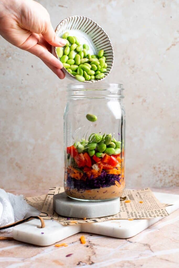Edamame being poured into a mason jar layered with cabbage and carrots