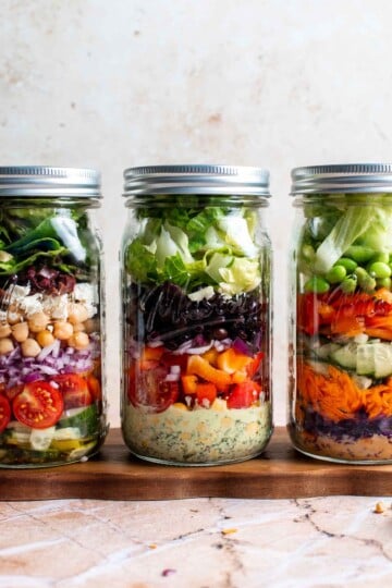 Three mason jar salads lined up showing colorful layered ingredients, including Mediterranean, Southwest, and Asian peanut variations