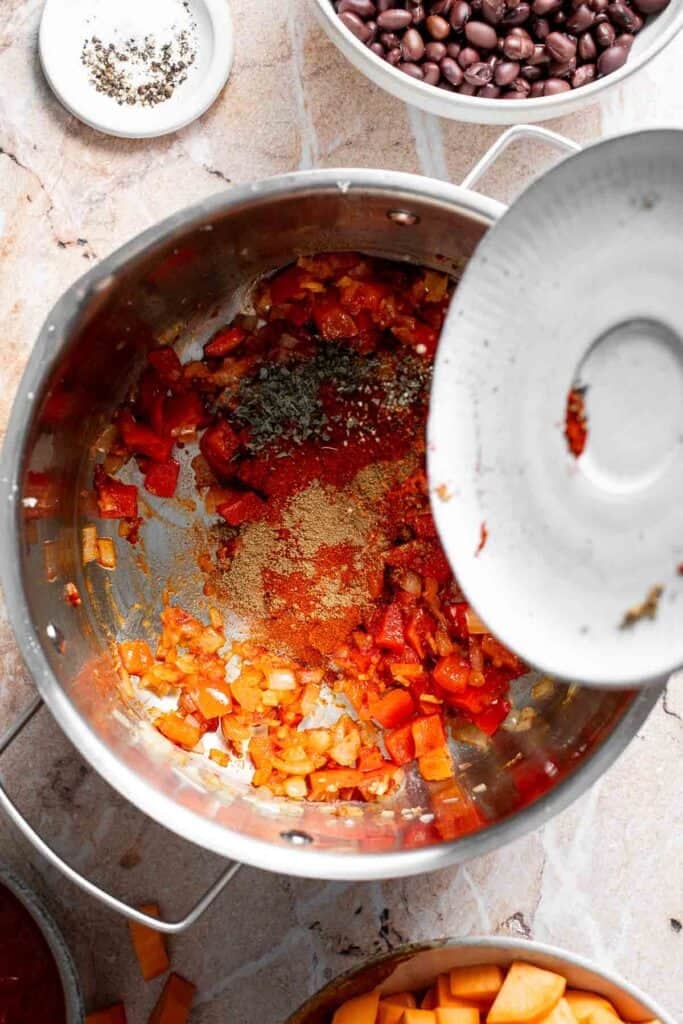 Adding spices including chili powder, cumin, smoked paprika, oregano, and coriander to the pot of veggies.