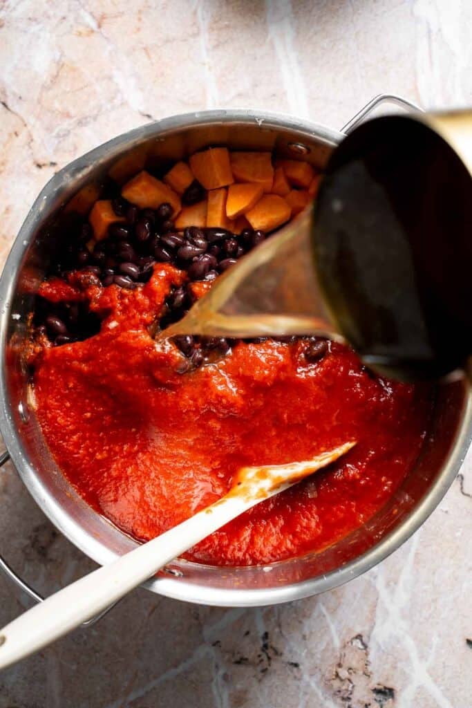 Adding sweet potatoes, diced tomatoes, tomato sauce, black beans, and vegetable broth to pot of veggies
