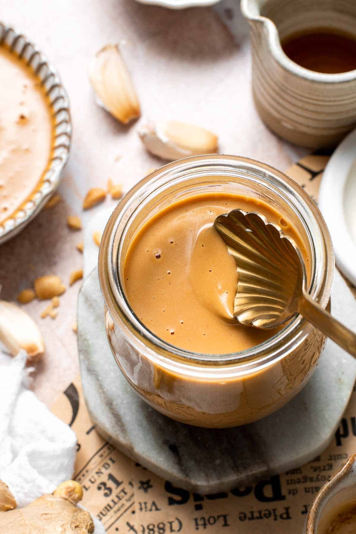 Scooping Thai peanut dressing from a glass jar surrounded by peanuts and garlic
