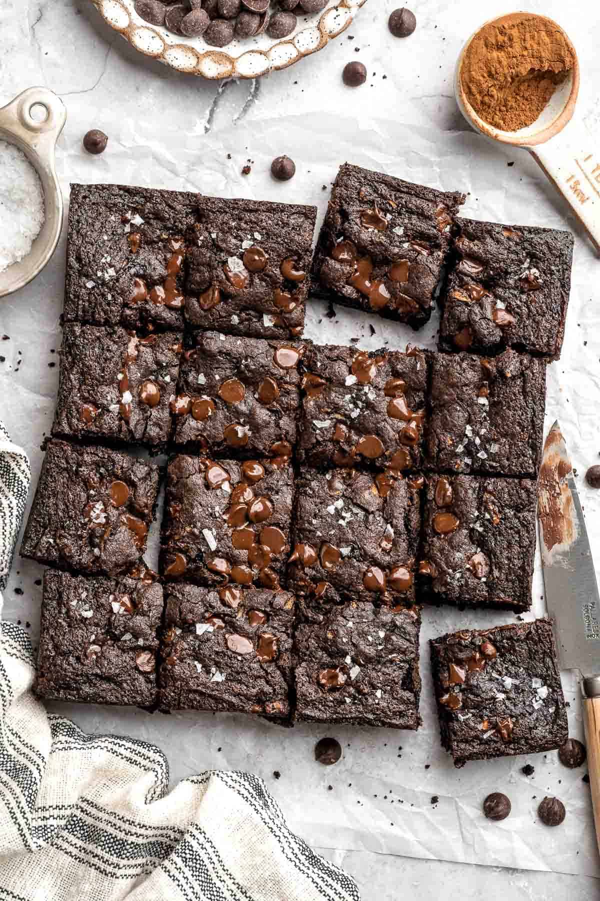 Overhead view of sliced zucchini brownies arranged on parchment paper surrounded by baking ingredients