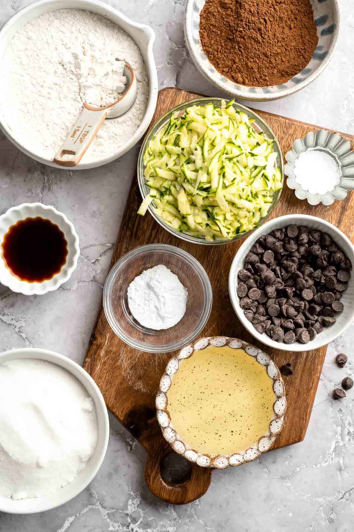 Ingredients for fudgy zucchini brownies including flour, cocoa powder, shredded zucchini, sugar, and chocolate chips arranged on a wood board
