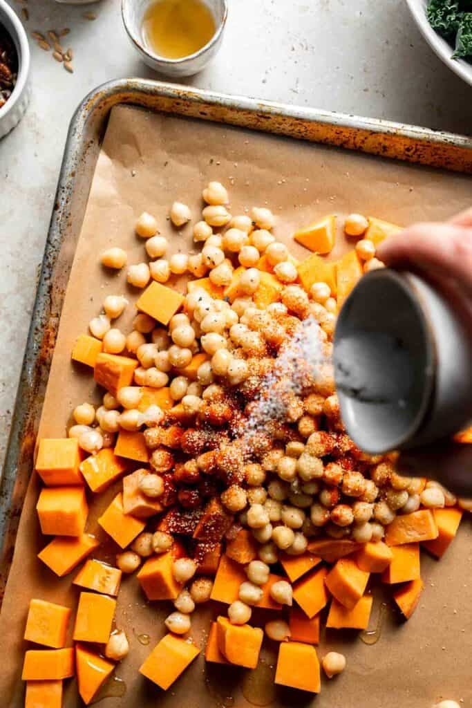 Salt and pepper being added over sweet potatoes and chickpeas on a baking sheet