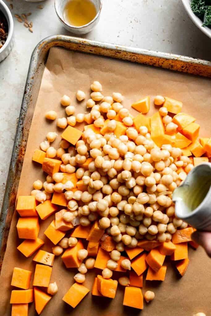 Olive oil being poured over sweet potatoes and chickpeas on a baking sheet