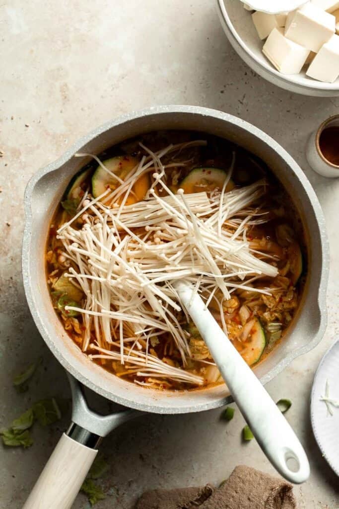 Enoki mushrooms being added to a pot of broth and vegetables