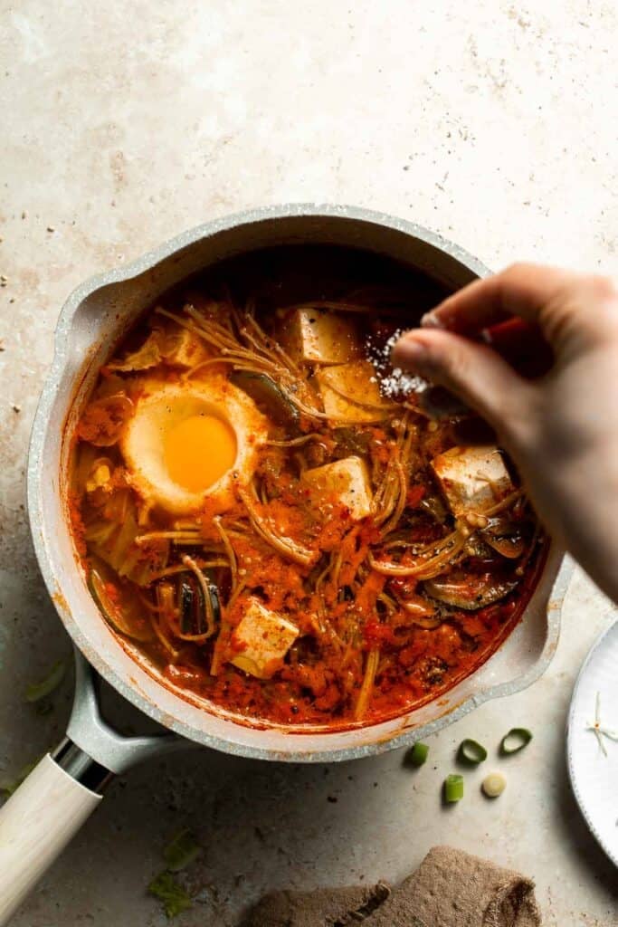 A hand adding sesame seeds to a pot of Korean tofu soup