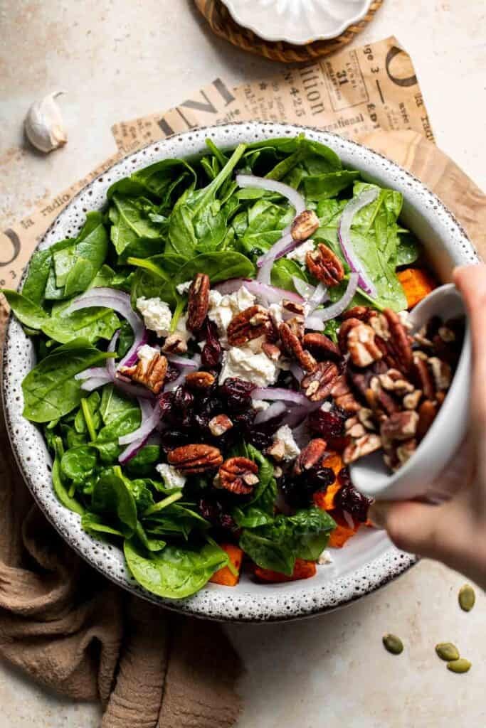 Adding pecans and feta cheese to bowl of baby spinach and roasted sweet potatoes