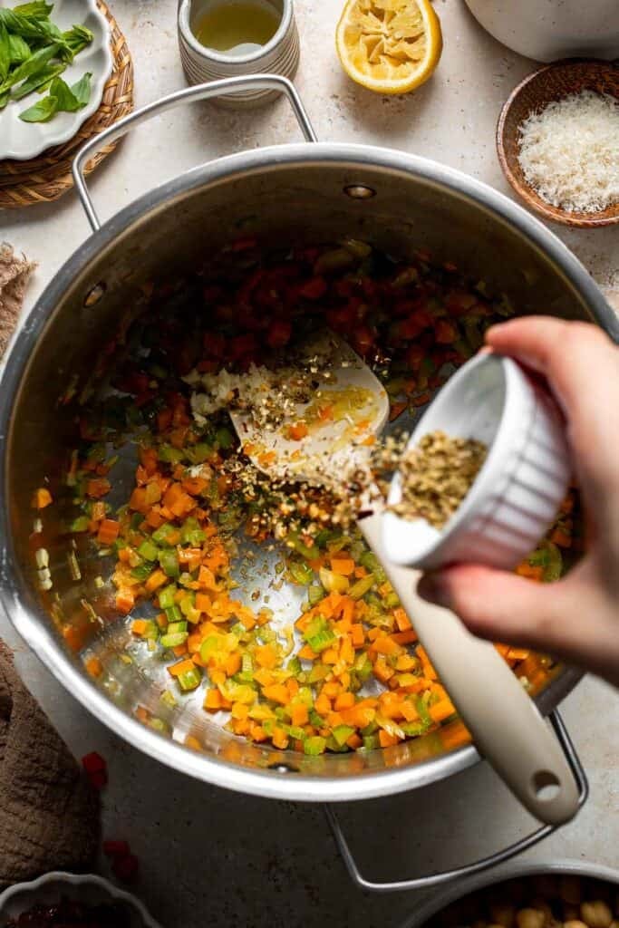 Dried oregano and spices being added to sautéed vegetables in a pot