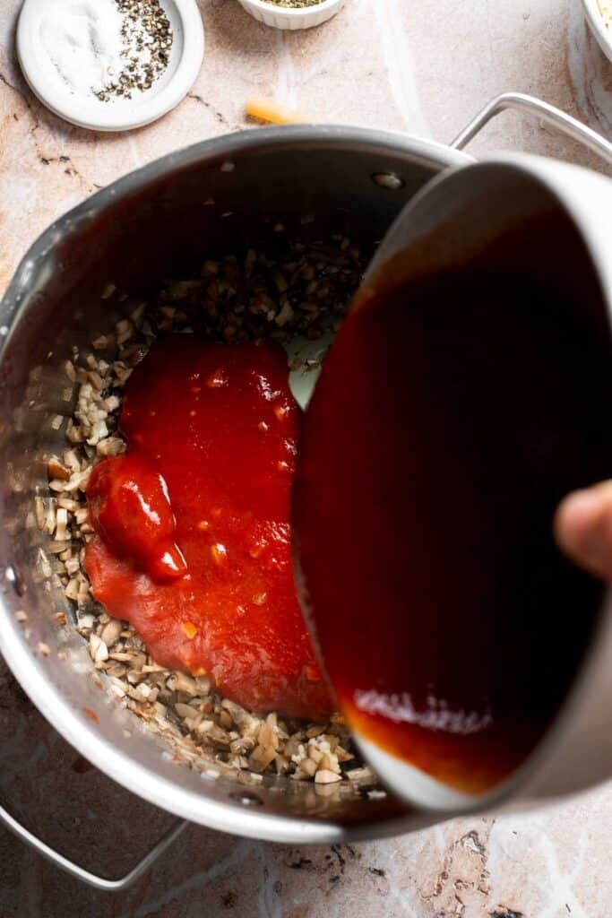 Marinara sauce being poured into a pot with cooked mushrooms and onion