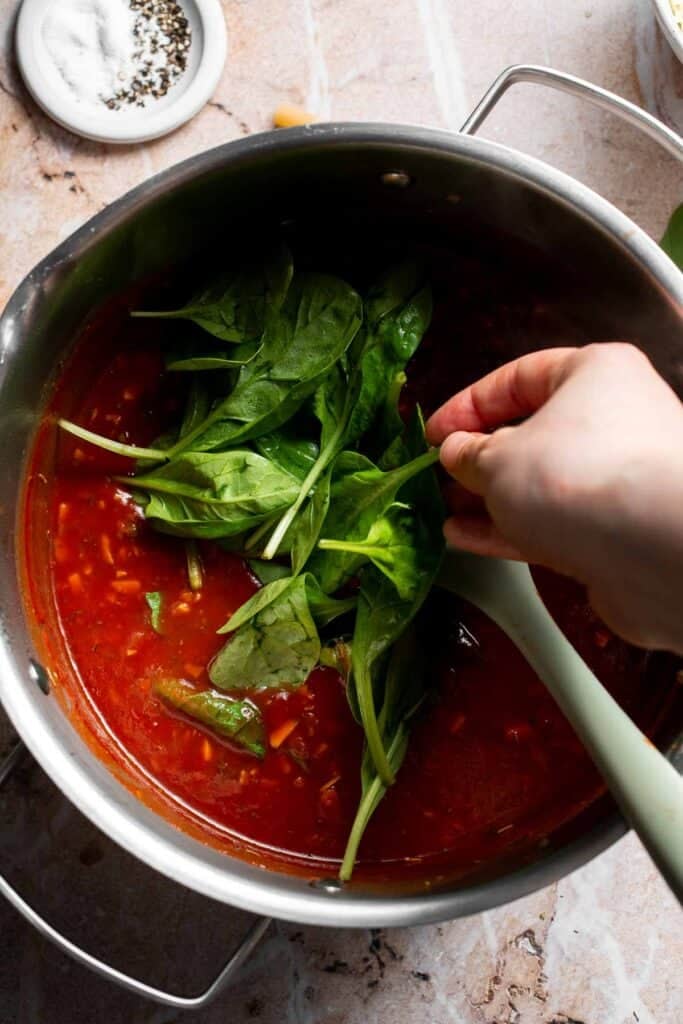 A handful of spinach being stirred into marinara sauce in a pot
