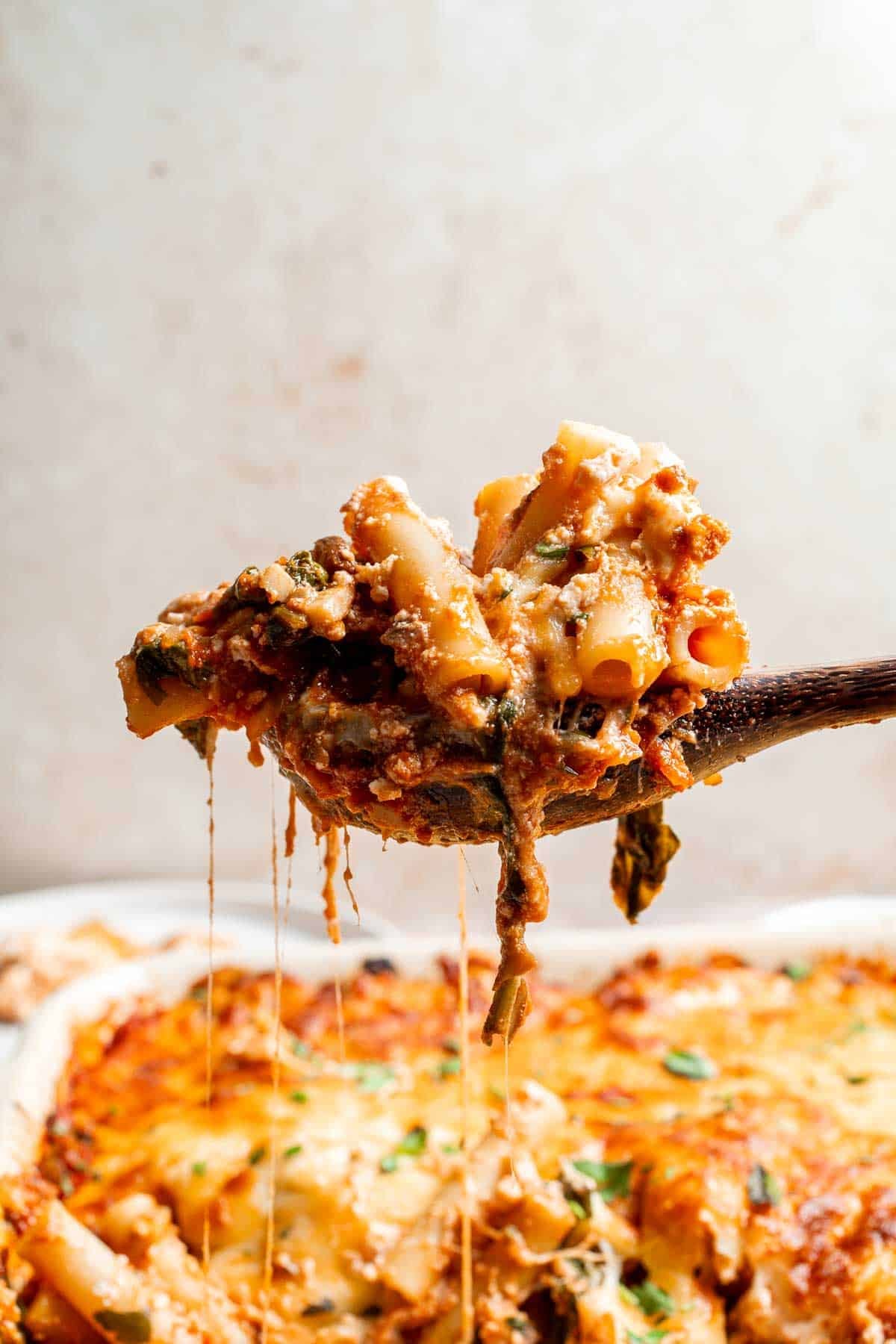 A serving spoon lifting a cheesy portion of baked ziti from the casserole dish