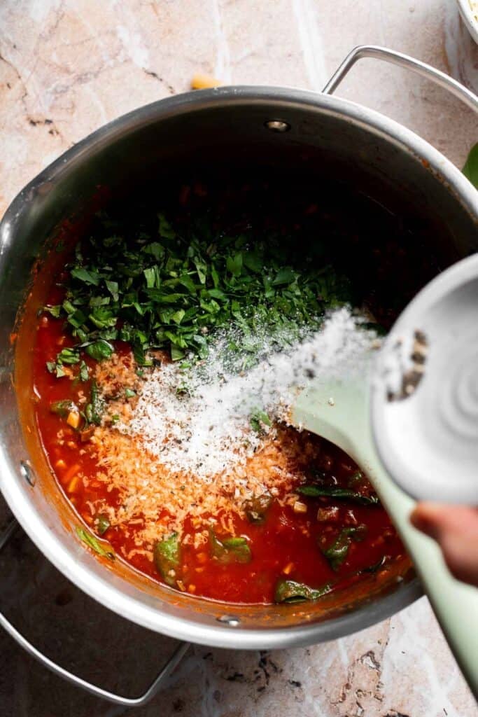Fresh parsley and basil being added to marinara sauce in a pot with salt and pepper