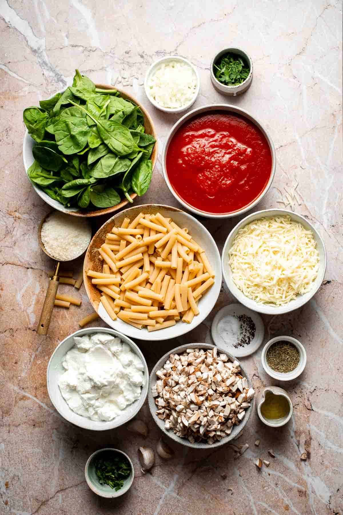 Ingredients for vegetarian baked ziti including spinach, marinara sauce, pasta, and cheese arranged in bowls